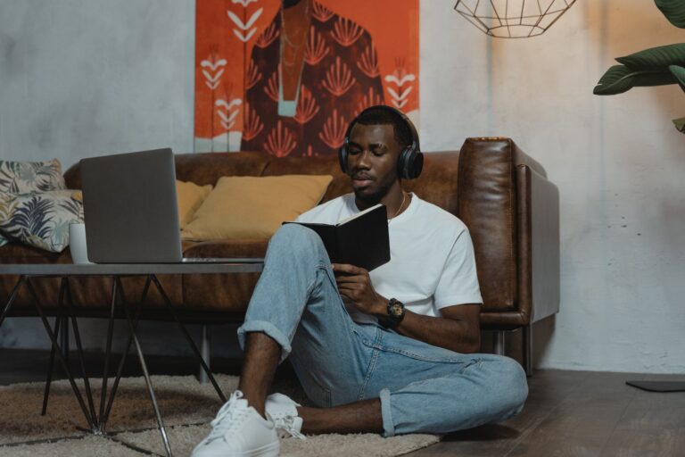 A man wearing headphones sitting on the floor in his flat reading a journal